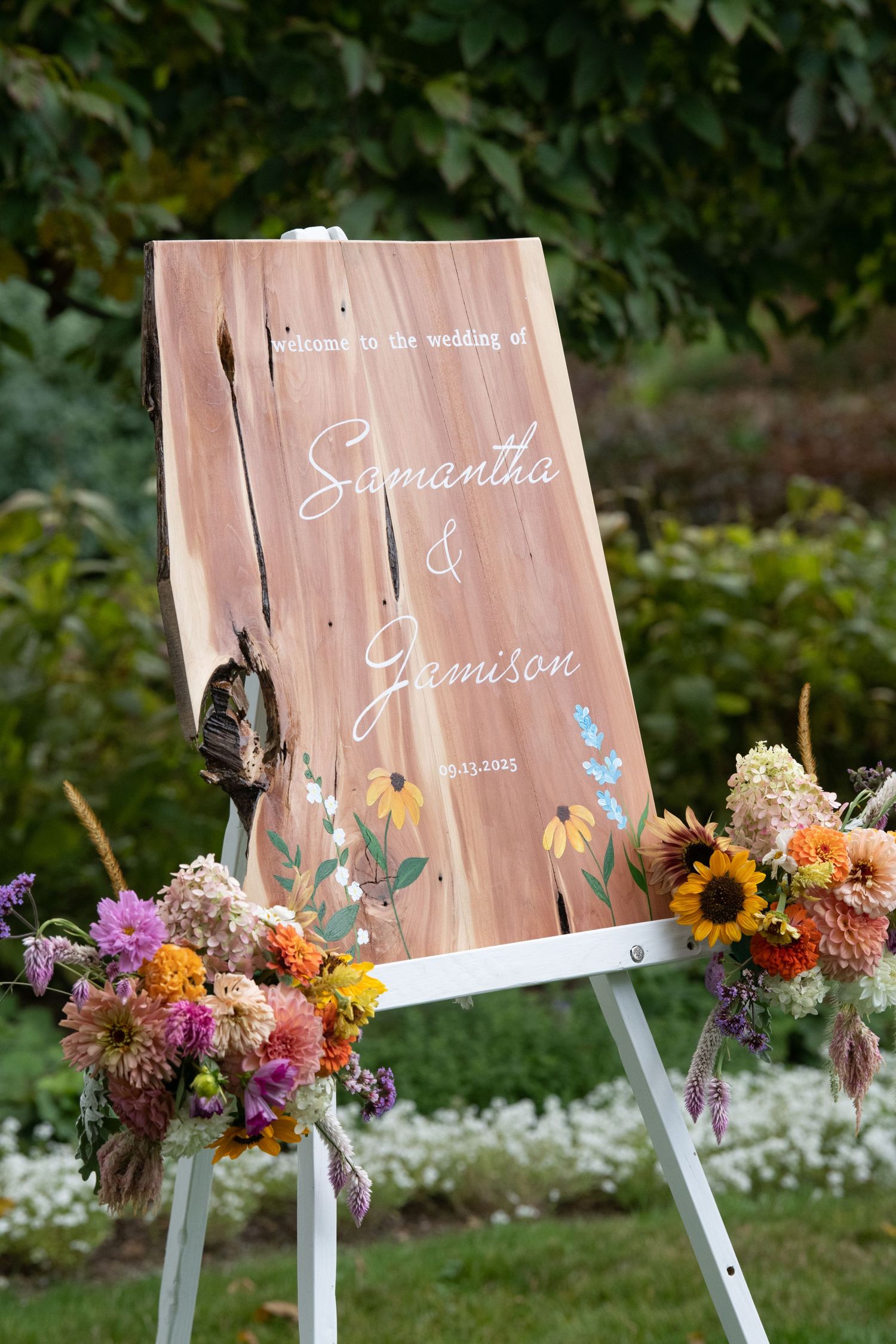 Wooden wedding sign decorated with colorful wildflowers and greenery displayed in an outdoor garden setting.