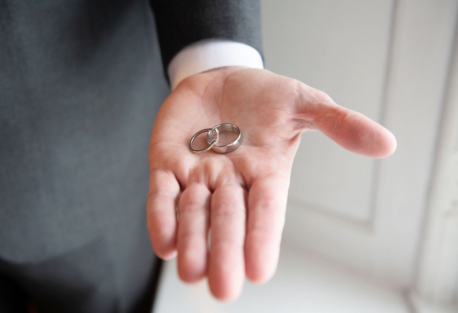 Two silver wedding rings rest in the palm of an outstretched hand wearing a white dress shirt sleeve.