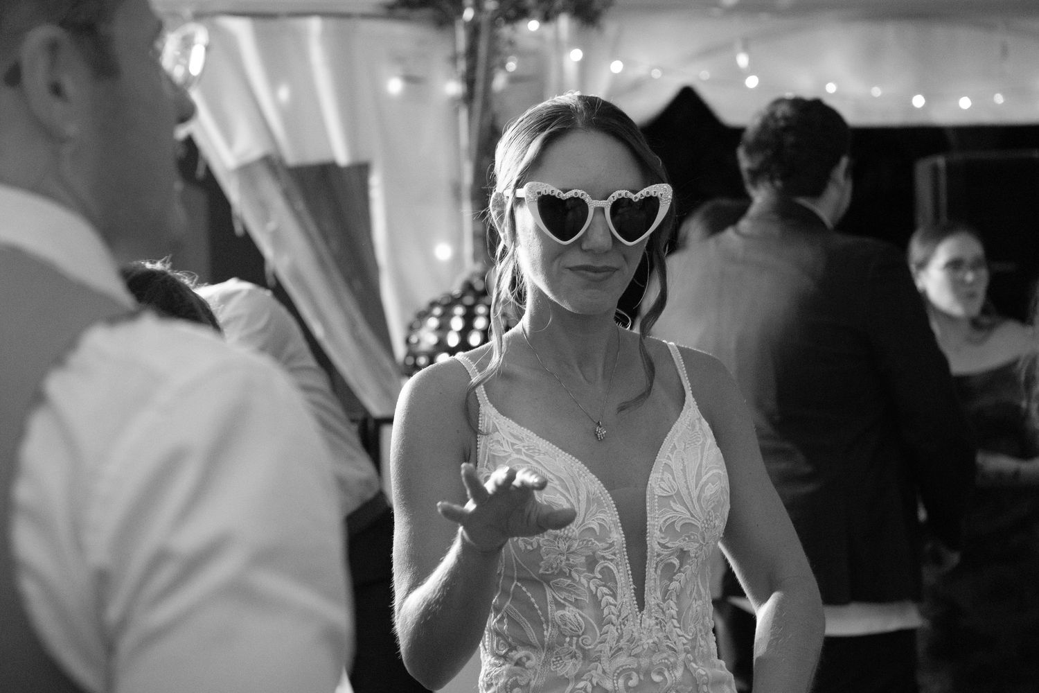 Wedding guest in sunglasses and white dress enjoying reception under string lights.
