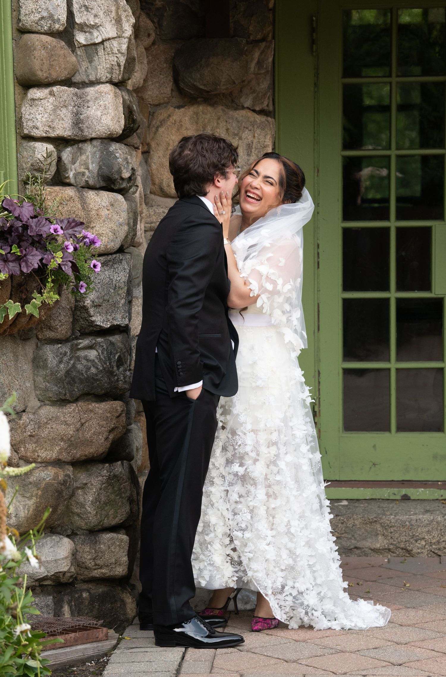 A couple in wedding attire share intimate moments against a rustic stone wall with green windows and purple flowers.