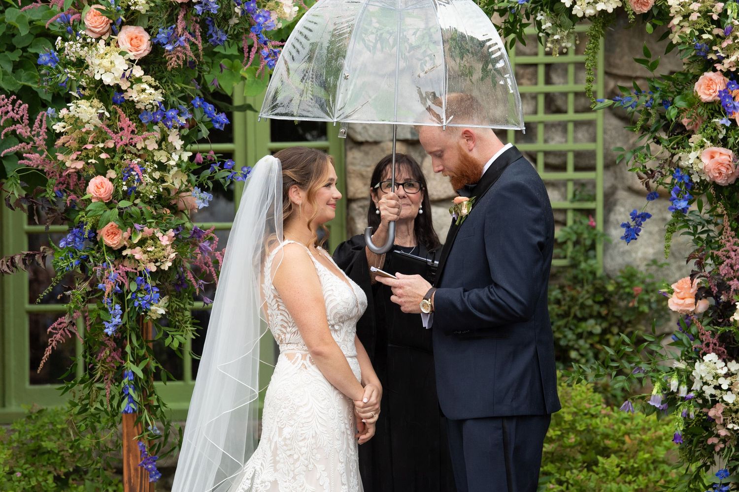 Wedding ceremony under floral arch with pink roses and blue flowers in a garden setting.