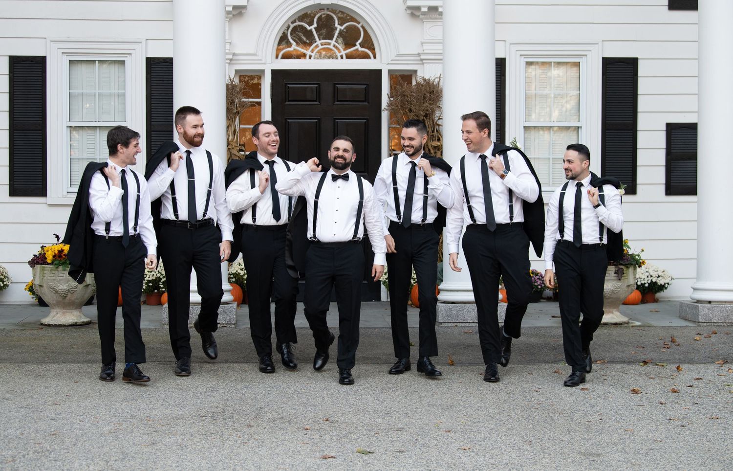 Groomsmen in black and white attire walk together outside a classic white building with black shutters.