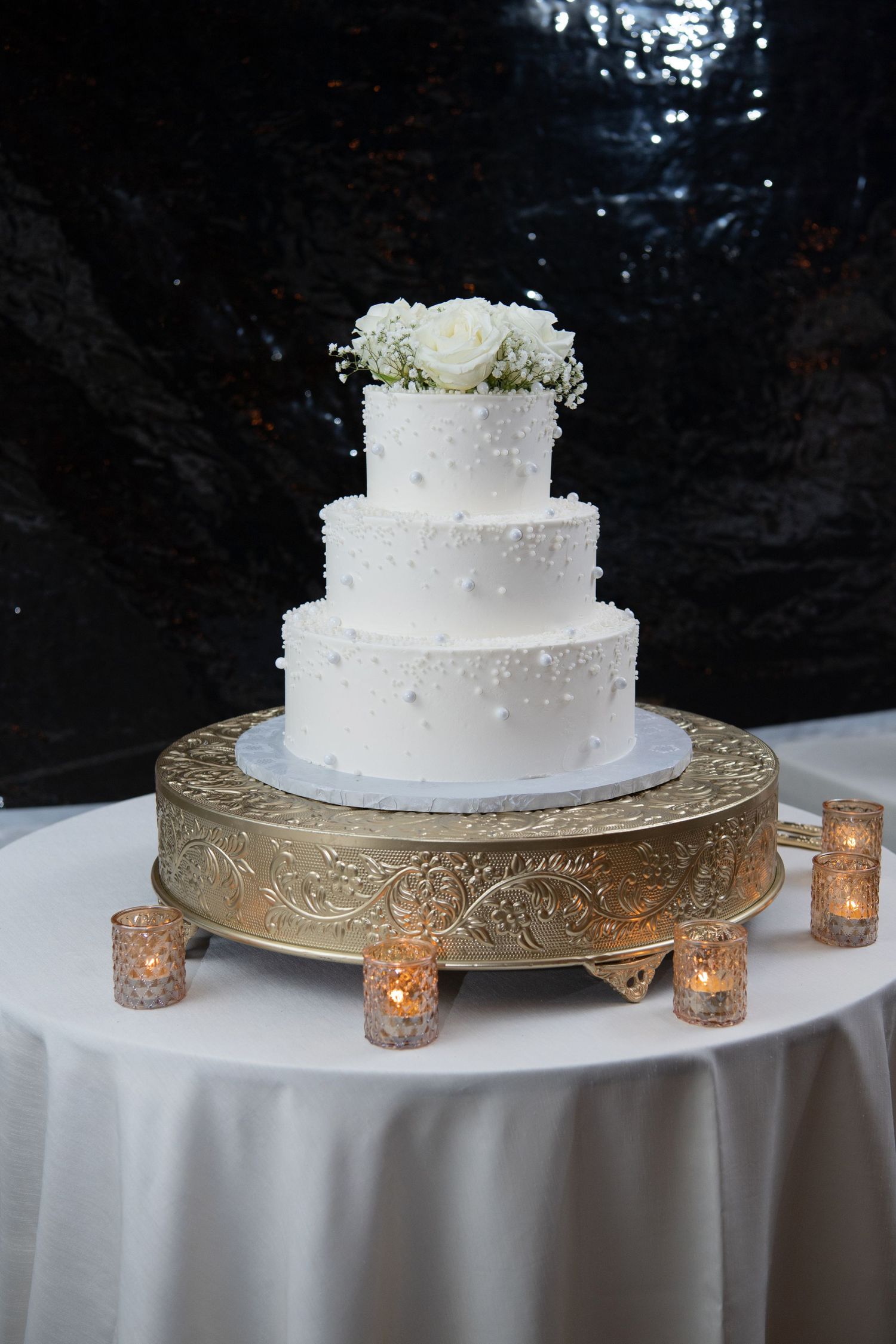 Elegant three-tier white wedding cake displayed on ornate gold stand with glowing votive candles on white tablecloth.
