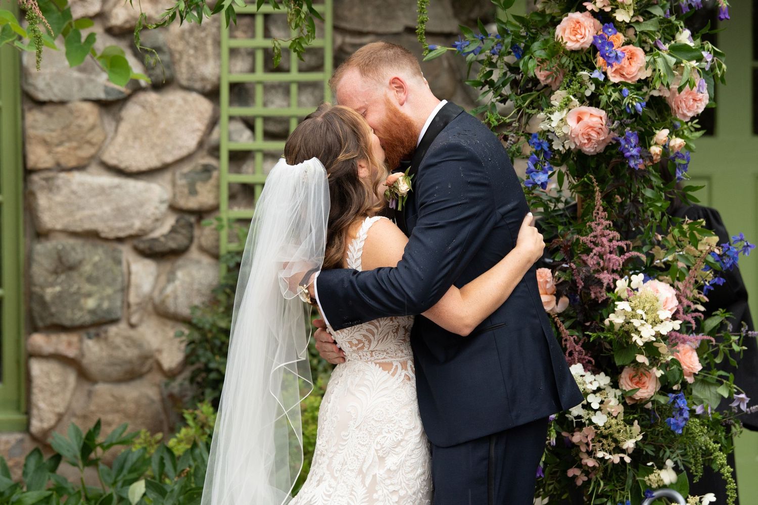 A romantic wedding kiss moment captured beside a stone wall adorned with climbing roses.