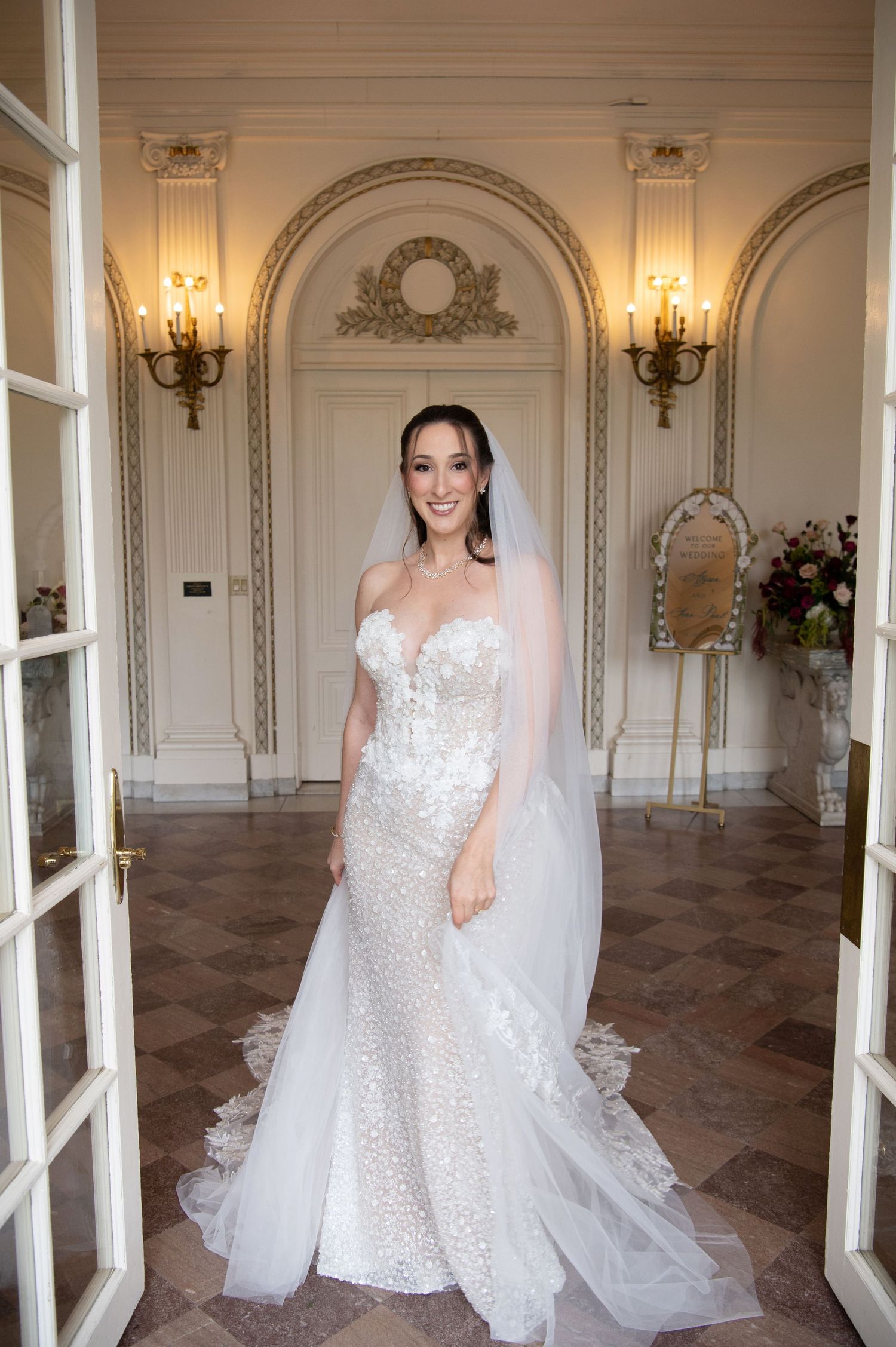 Bride in a beaded strapless mermaid wedding gown with veil poses in an elegant white ornate venue.