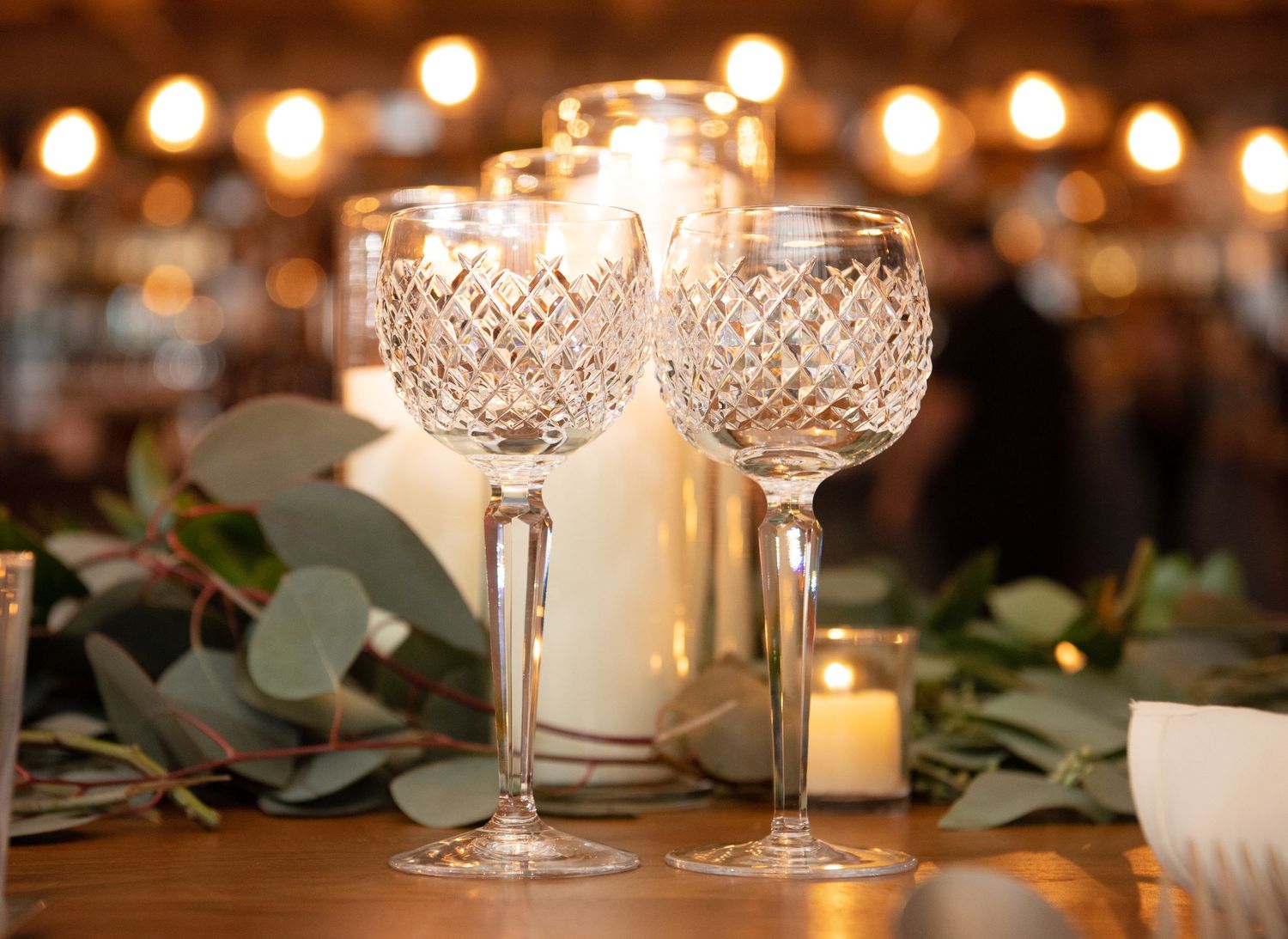 Crystal wine glasses and candles create an elegant table setting with eucalyptus greenery and warm glowing lights.
