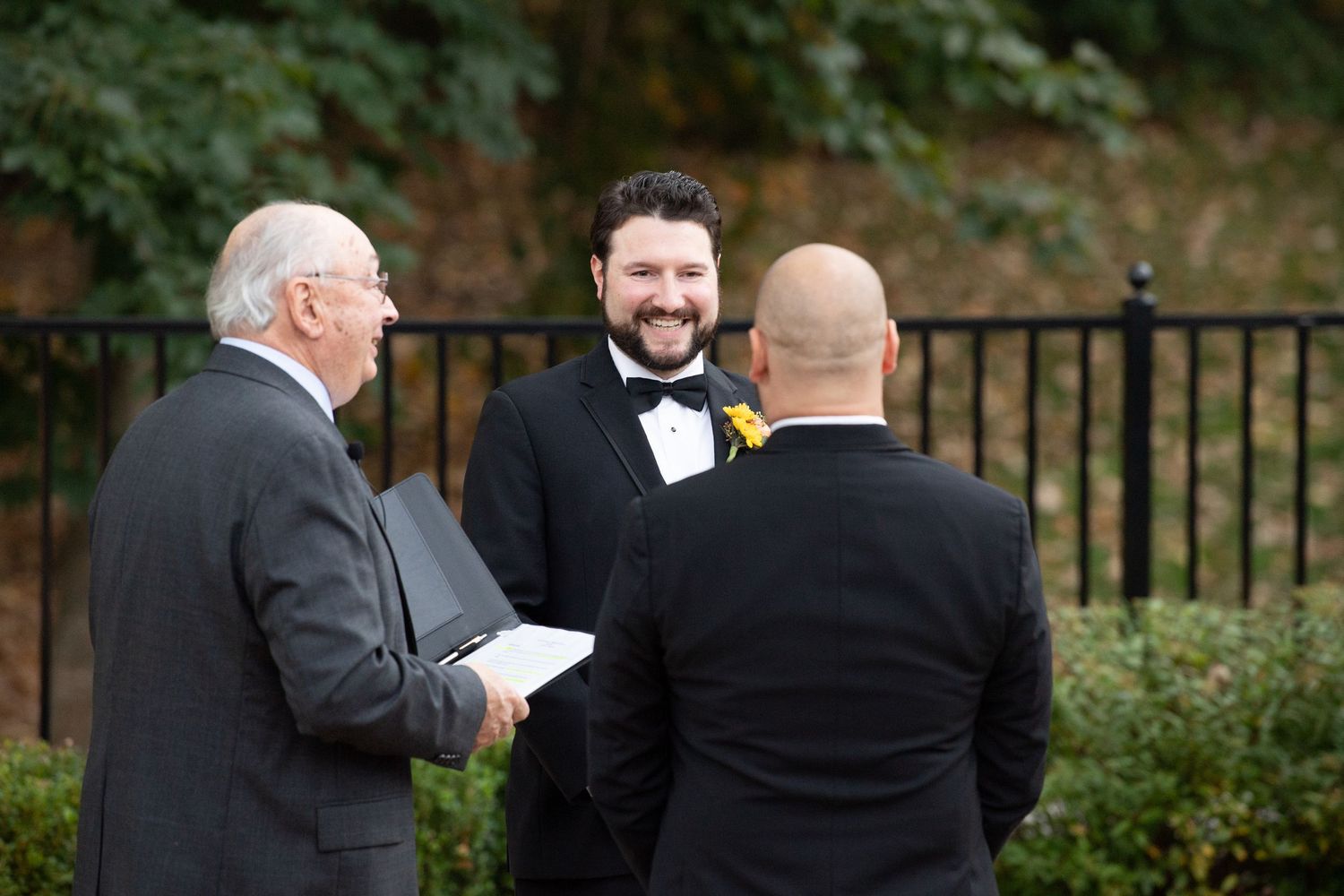 Two people stand together during an outdoor wedding ceremony with trees and a black metal railing in the background.