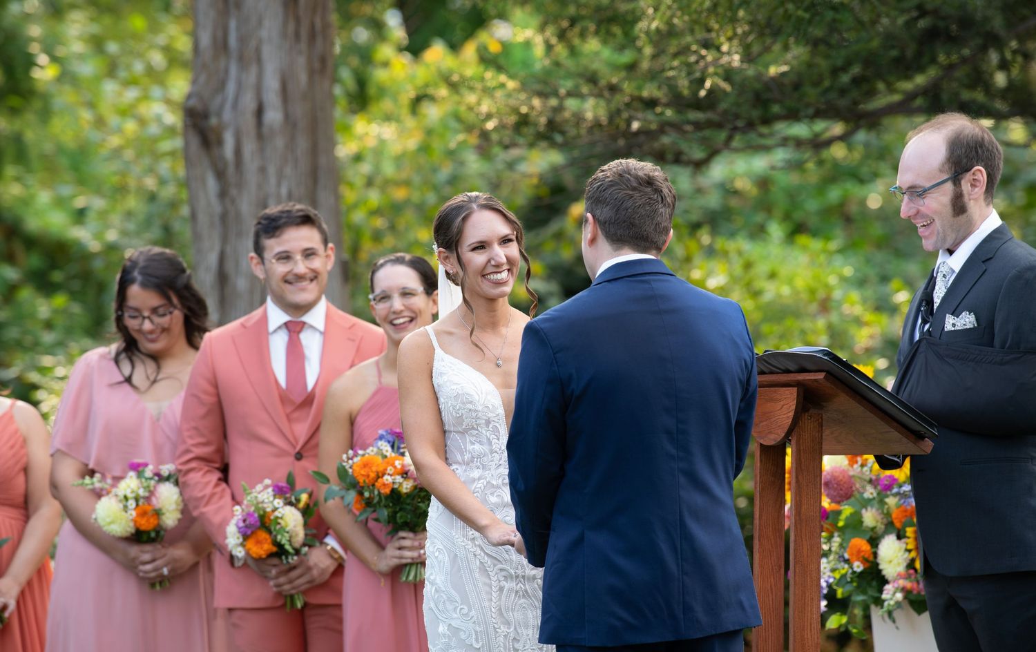 A wedding ceremony takes place outdoors with bridesmaids in coral dresses and guests seated on wooden benches.