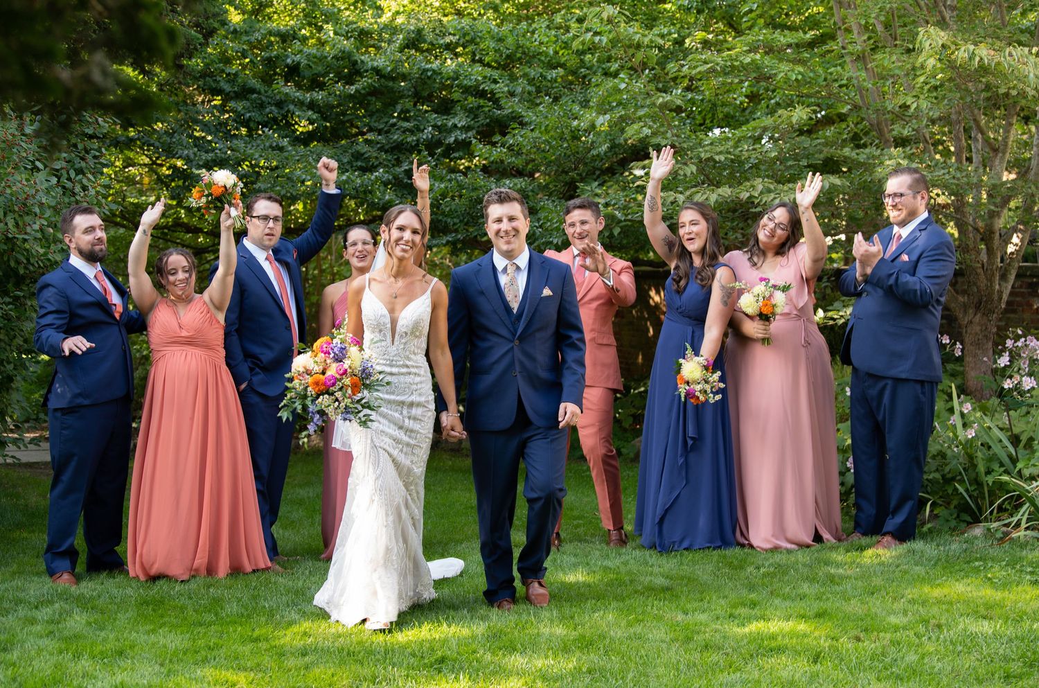 Wedding party in colorful attire poses on green lawn with bridesmaids in coral and pink dresses and groomsmen in navy suits.