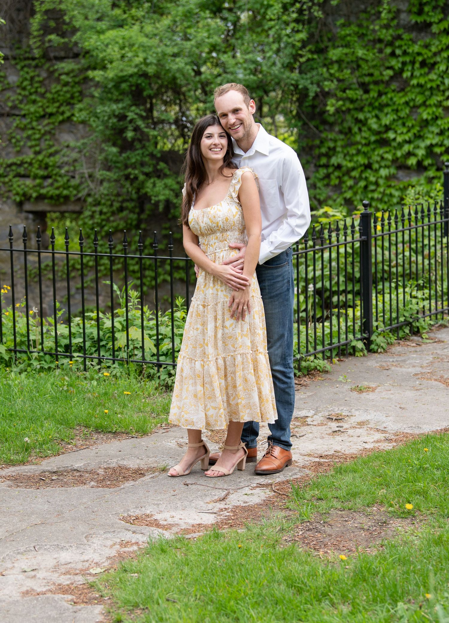 A couple shares romantic moments in a lush garden setting with an iron fence and stone pathway in golden hour light.