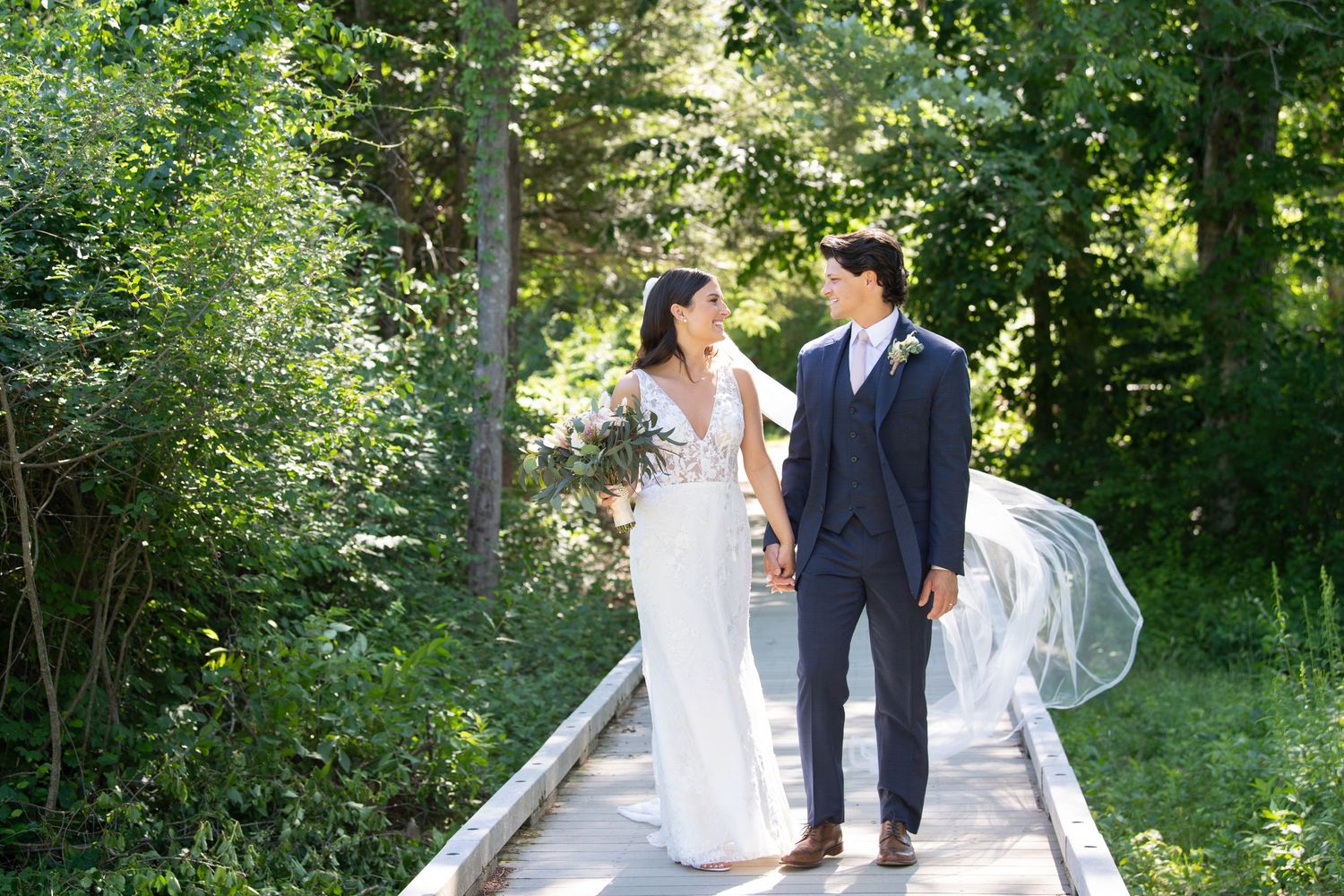A couple in wedding attire walks along a tree-lined path during an outdoor photo session.