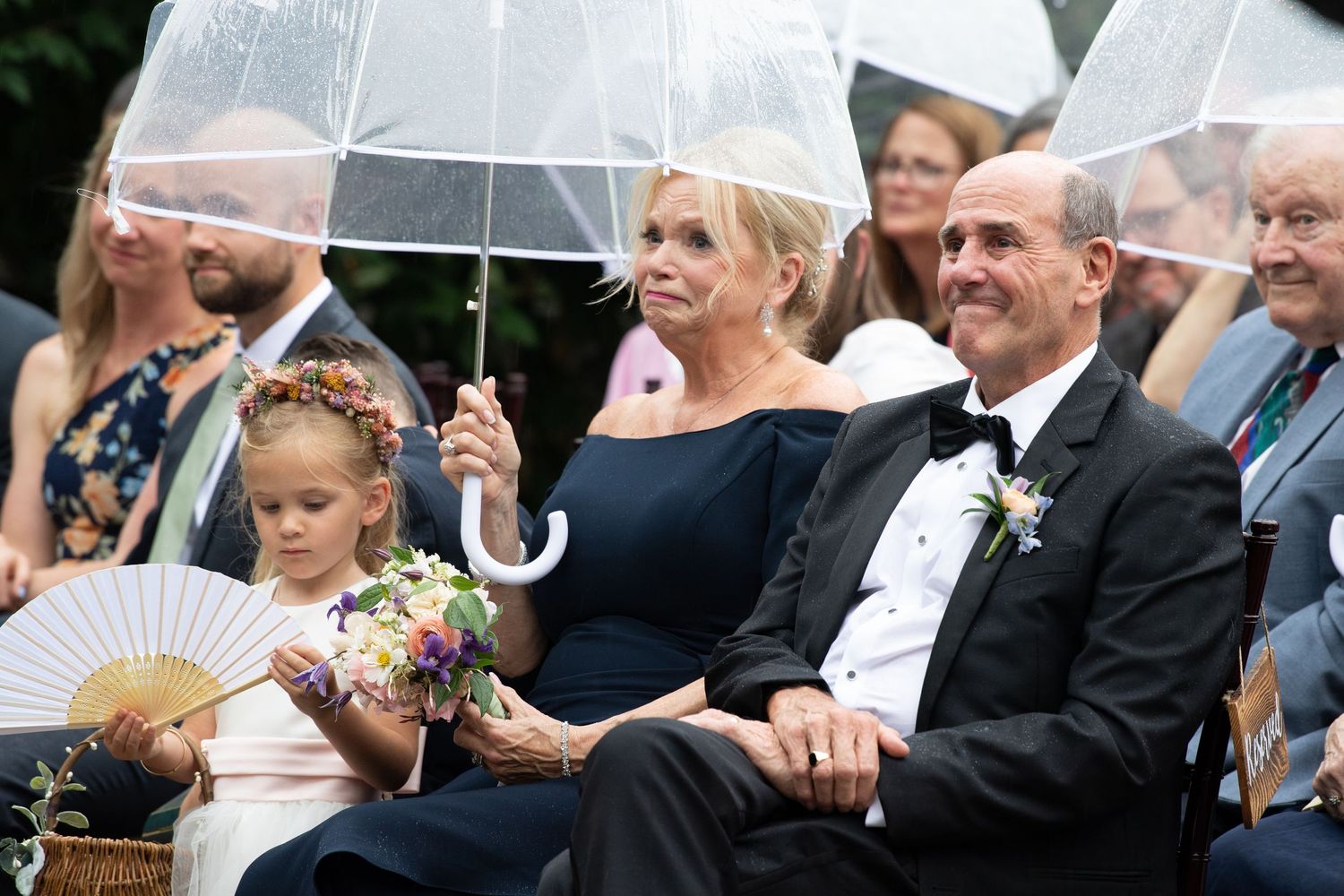 Wedding attendees sharing laughs while sheltered under clear umbrellas at an outdoor celebration.