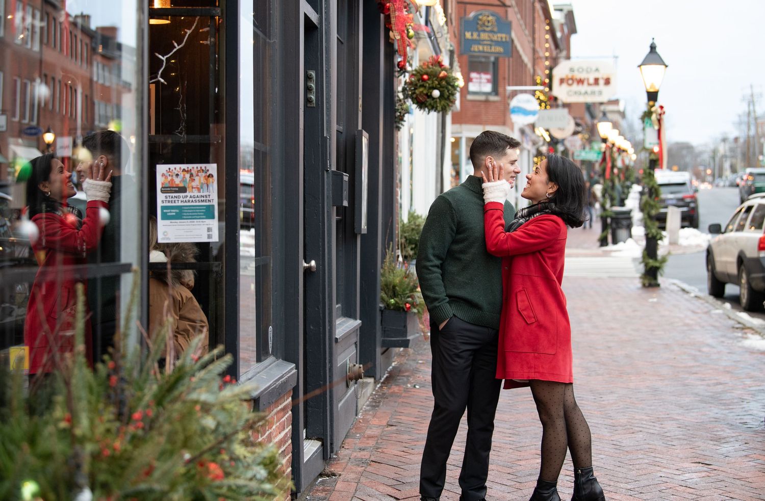 A couple in winter coats stands close together on a decorated brick sidewalk during the holiday season.