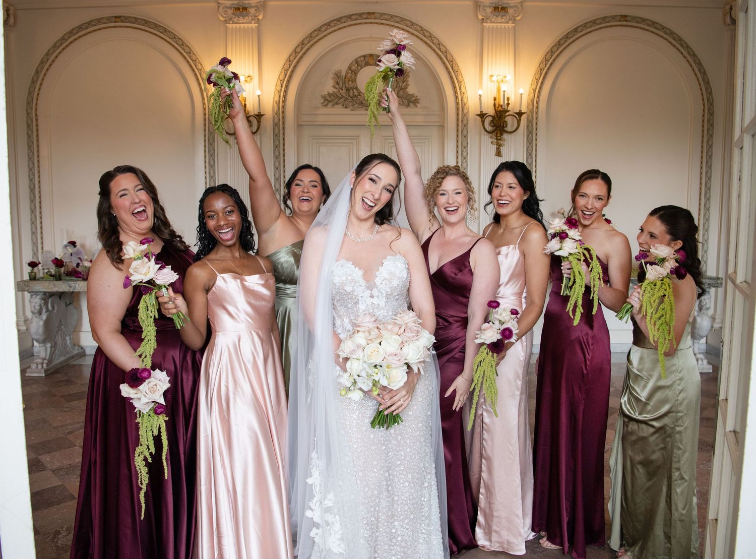 Bride in white lace gown with veil poses with six bridesmaids in mismatched burgundy and blush satin gowns holding bouquets.