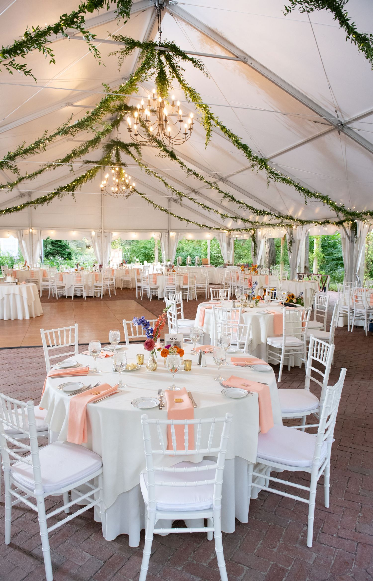 Elegant outdoor wedding reception under a white tent with green garland, chandeliers, white chiavari chairs, and peach napkins.
