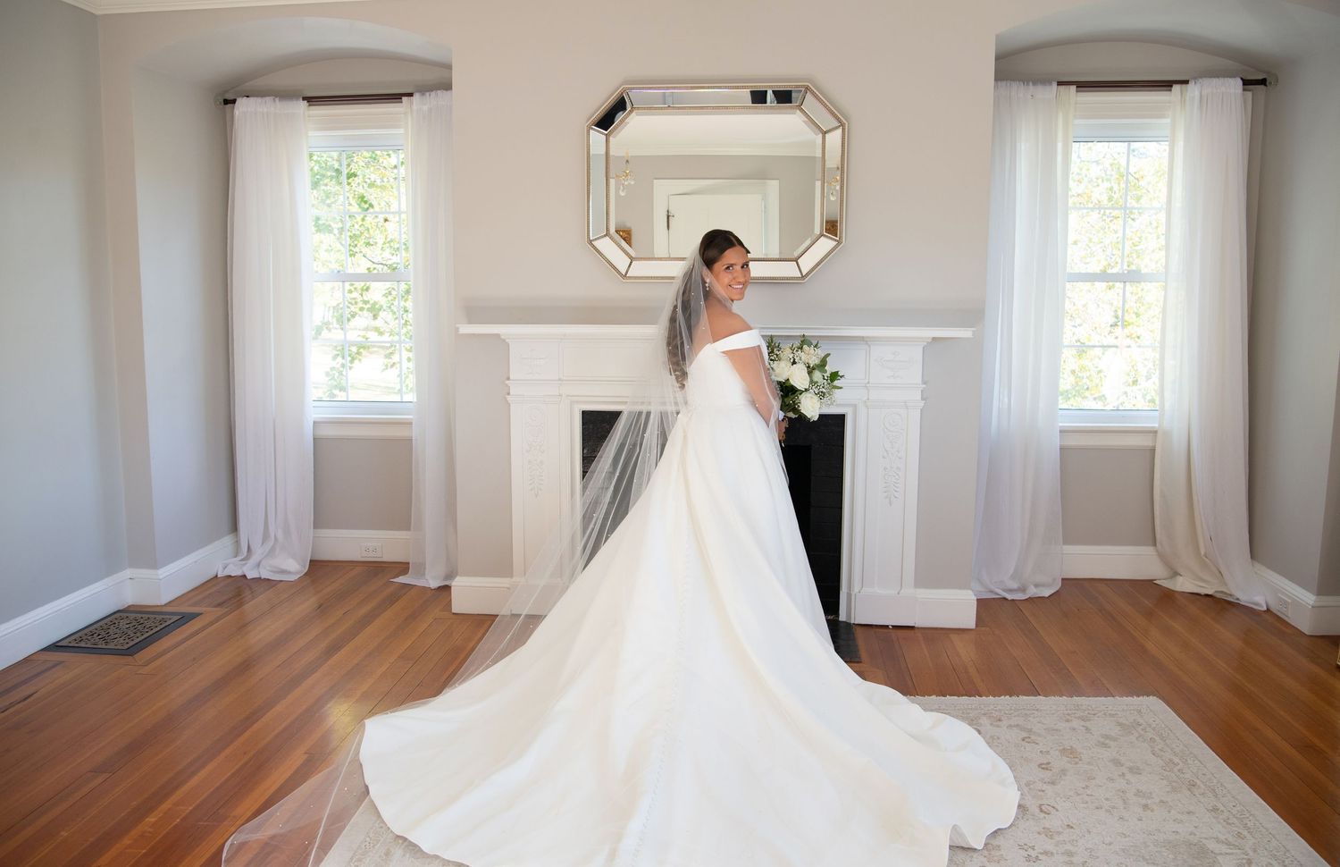 A wedding dress train flows across a hardwood floor in a bright white room with multiple windows.