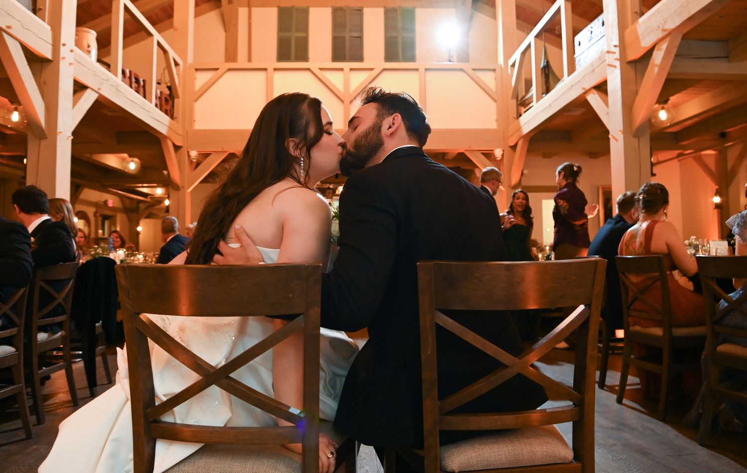 Wedding reception guests dance and celebrate in a rustic wooden barn venue illuminated with warm lighting.