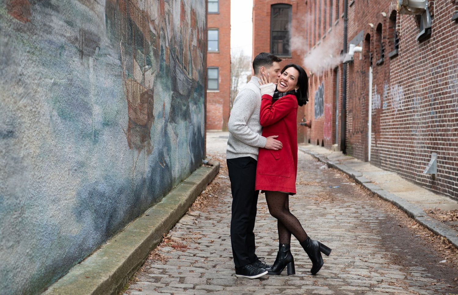 A couple embraces romantically on a cobblestone alley with brick buildings and a foggy atmosphere.