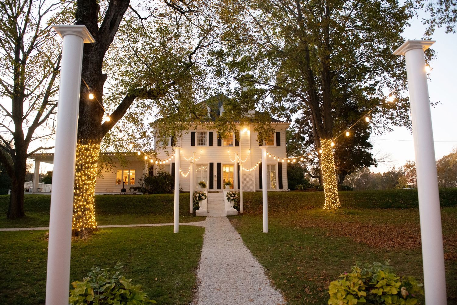 White columns line a walkway leading to a historic mansion surrounded by autumn trees at sunset.