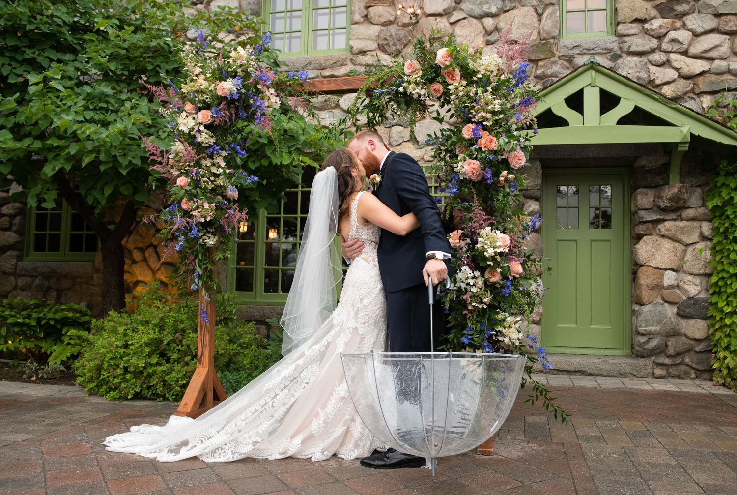 Couple embracing under a floral arch against a rustic stone building with a green door.