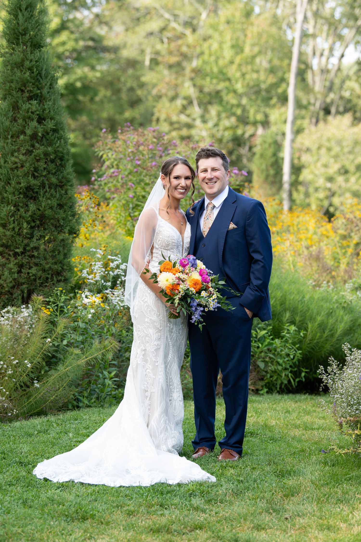 Couple standing in a lush garden with colorful summer flowers and evergreen trees.