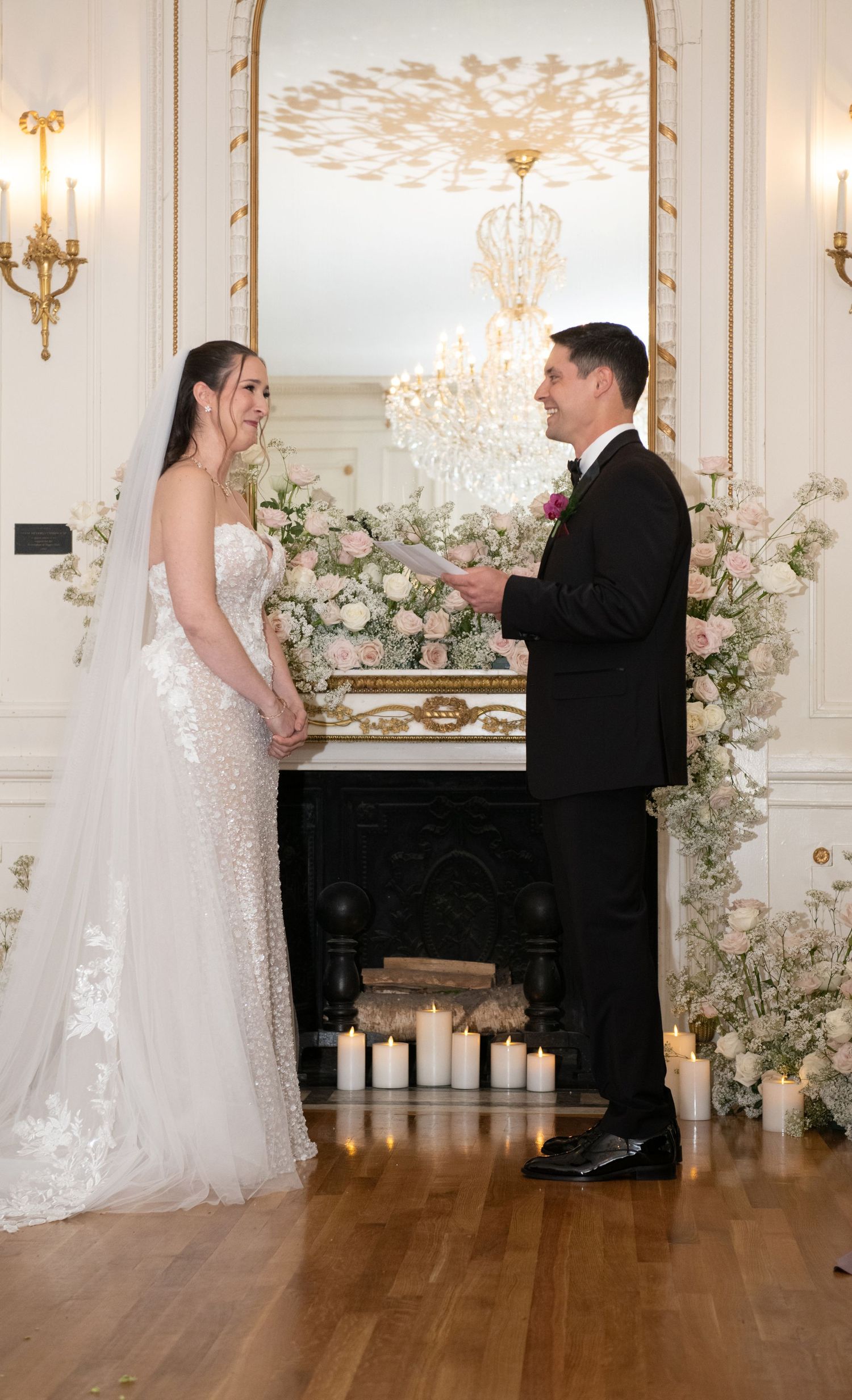 Bride and groom exchange vows by a floral fireplace with candles in an elegant white and gold ballroom.