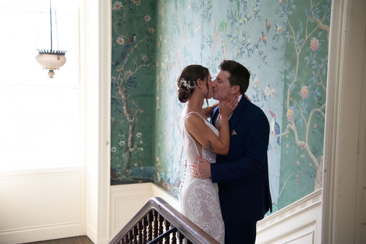 Bride and groom share a romantic kiss on a staircase landing with elegant green floral wallpaper backdrop.