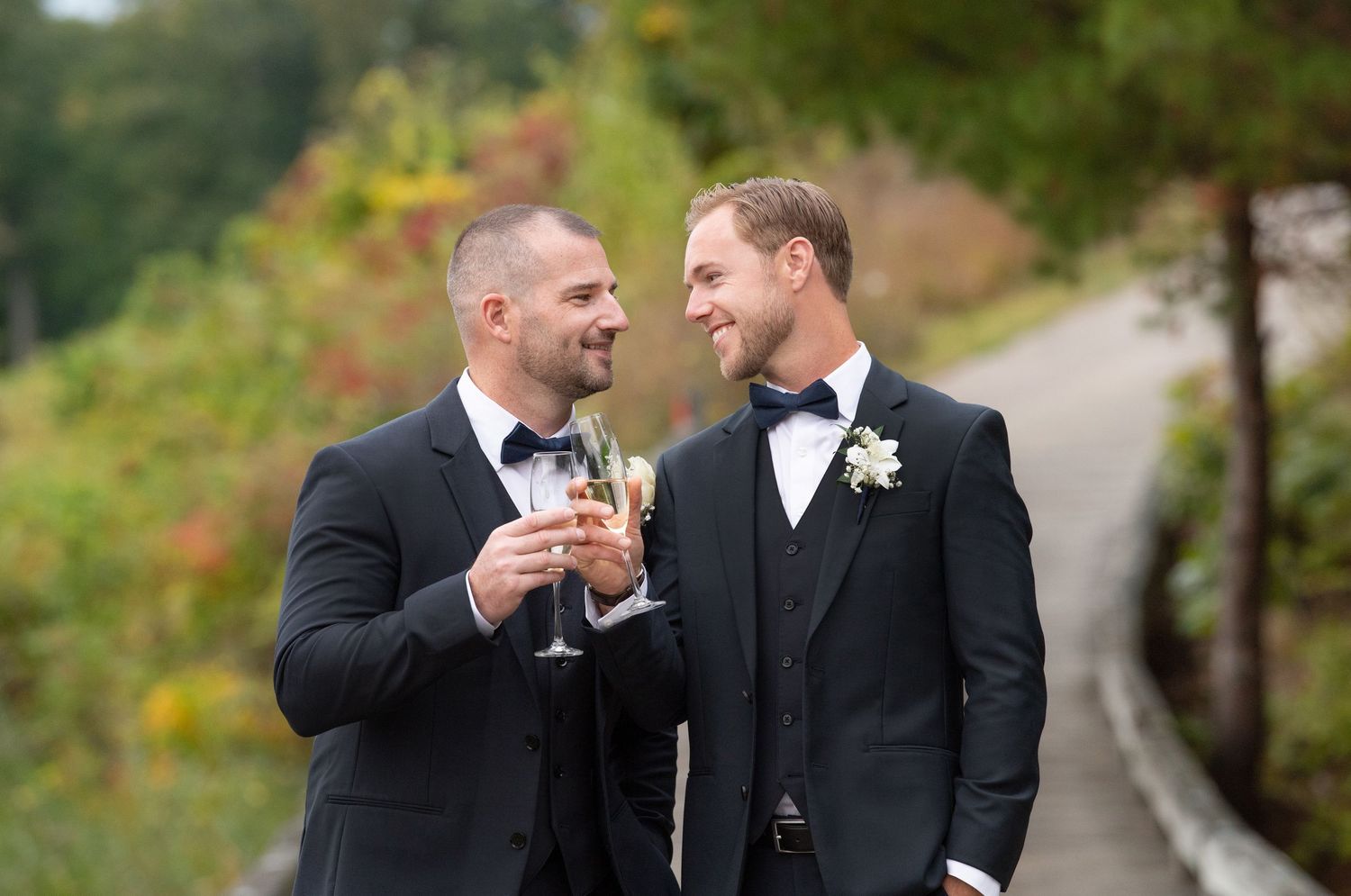 Two people in black suits laughing and toasting champagne glasses in an outdoor garden setting. LGBT wedding, Renaissance Haverhill wedding