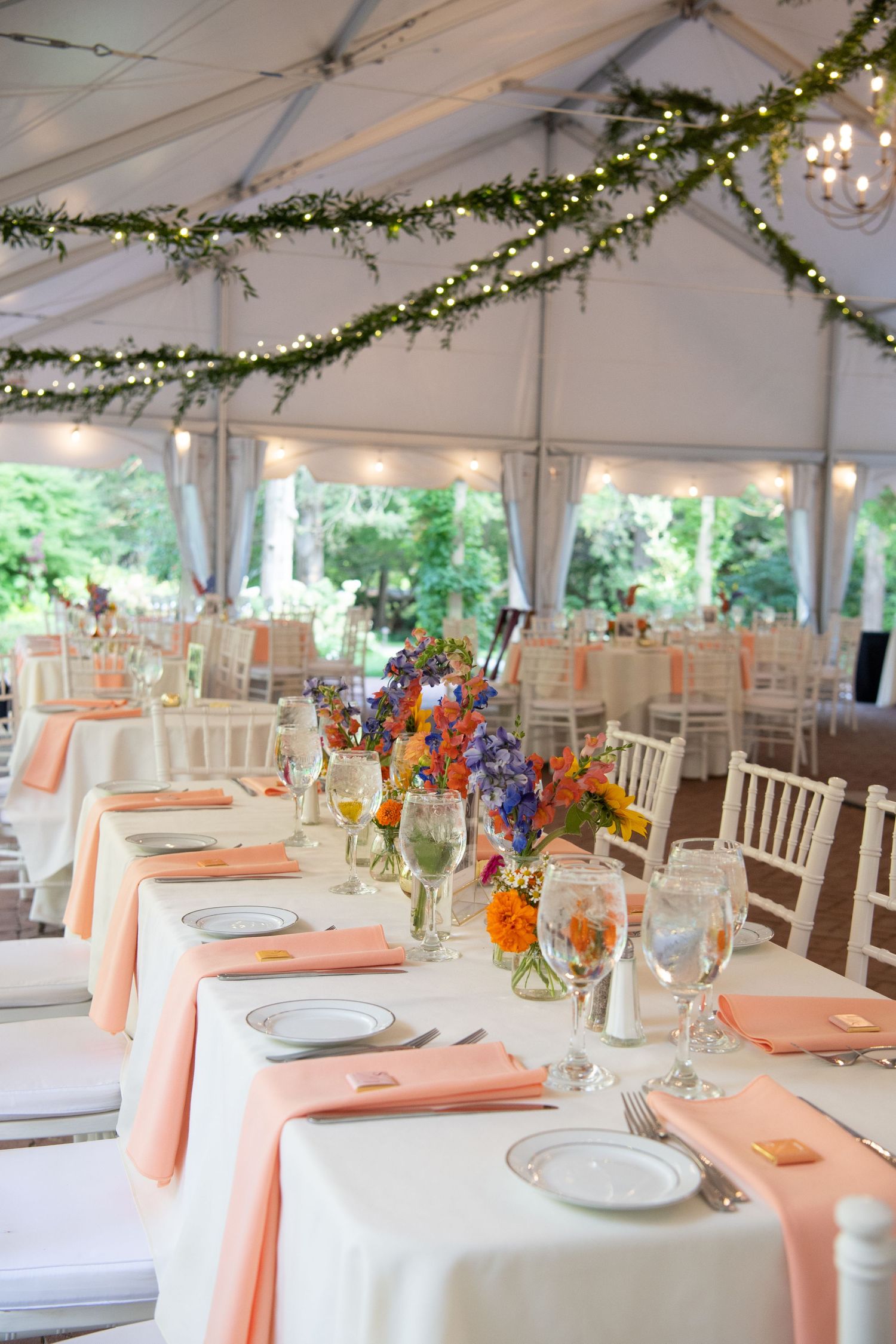 Wedding reception tent with elegant table settings featuring peach napkins and string lights overhead.