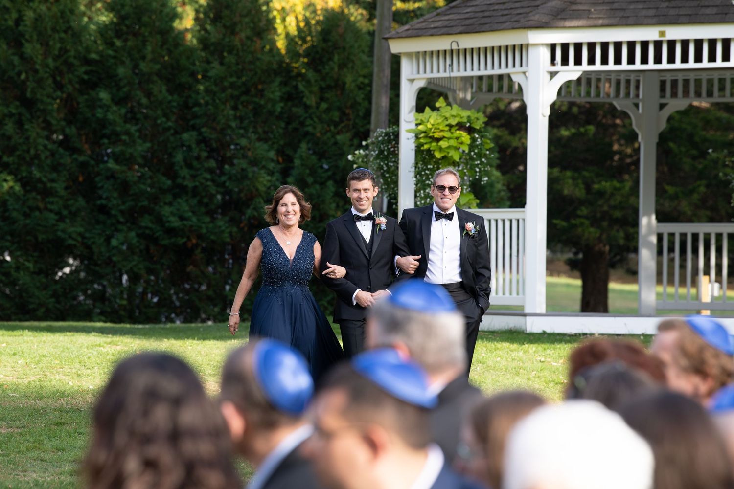 Wedding ceremony processional at an outdoor venue with white gazebo and seated guests.