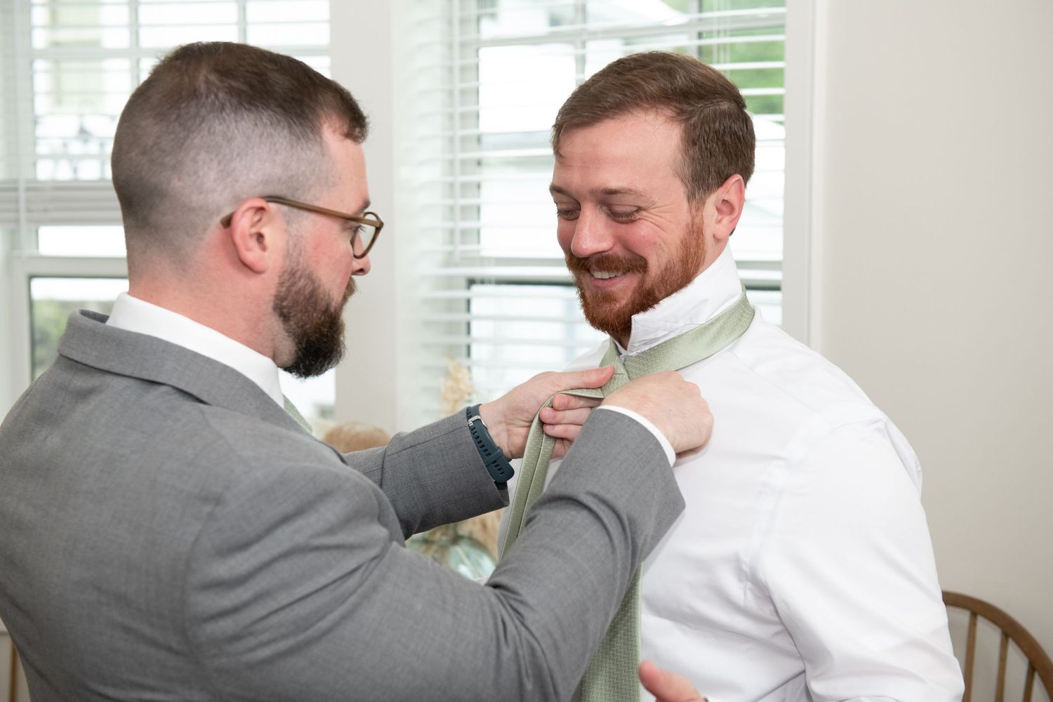 Two men in formal attire share a lighthearted moment while adjusting each other's bow ties.