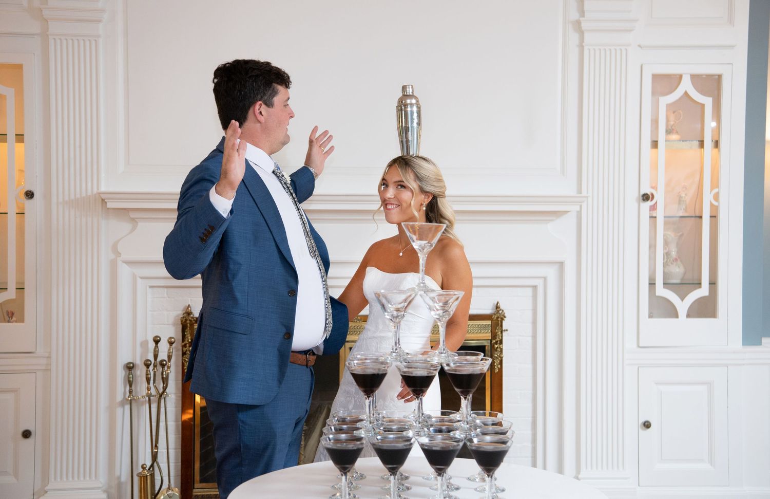 Two people stand next to a champagne tower with red wine glasses in an elegant white room with ornate molding.