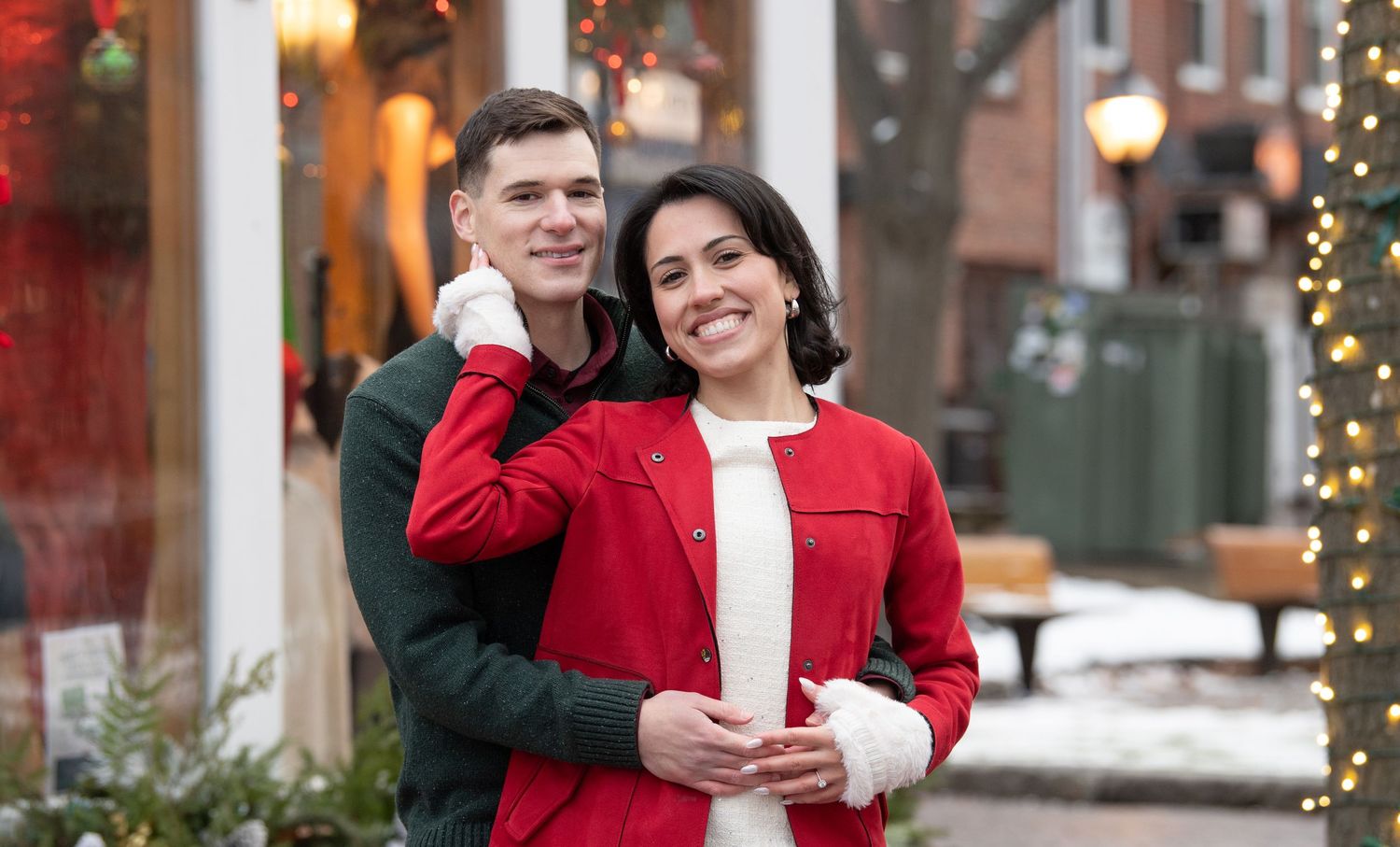 A couple embraces outdoors during winter, with the person in front wearing a festive red sweater near holiday decorations.