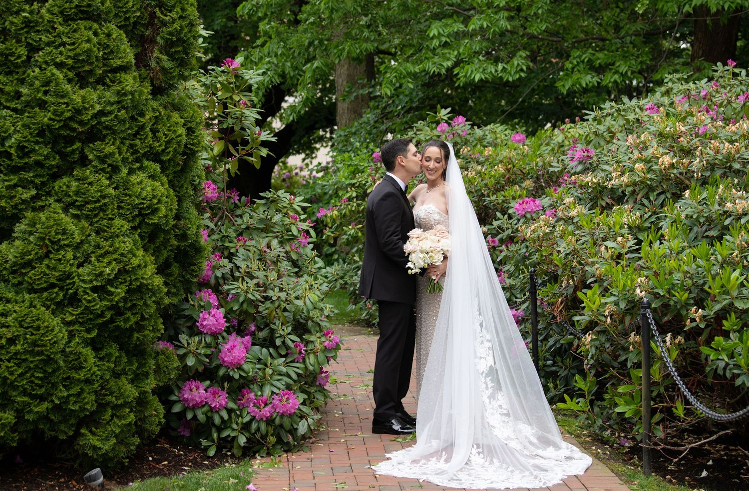 A bride and groom share a romantic kiss in a lush garden surrounded by blooming pink and purple flowers.