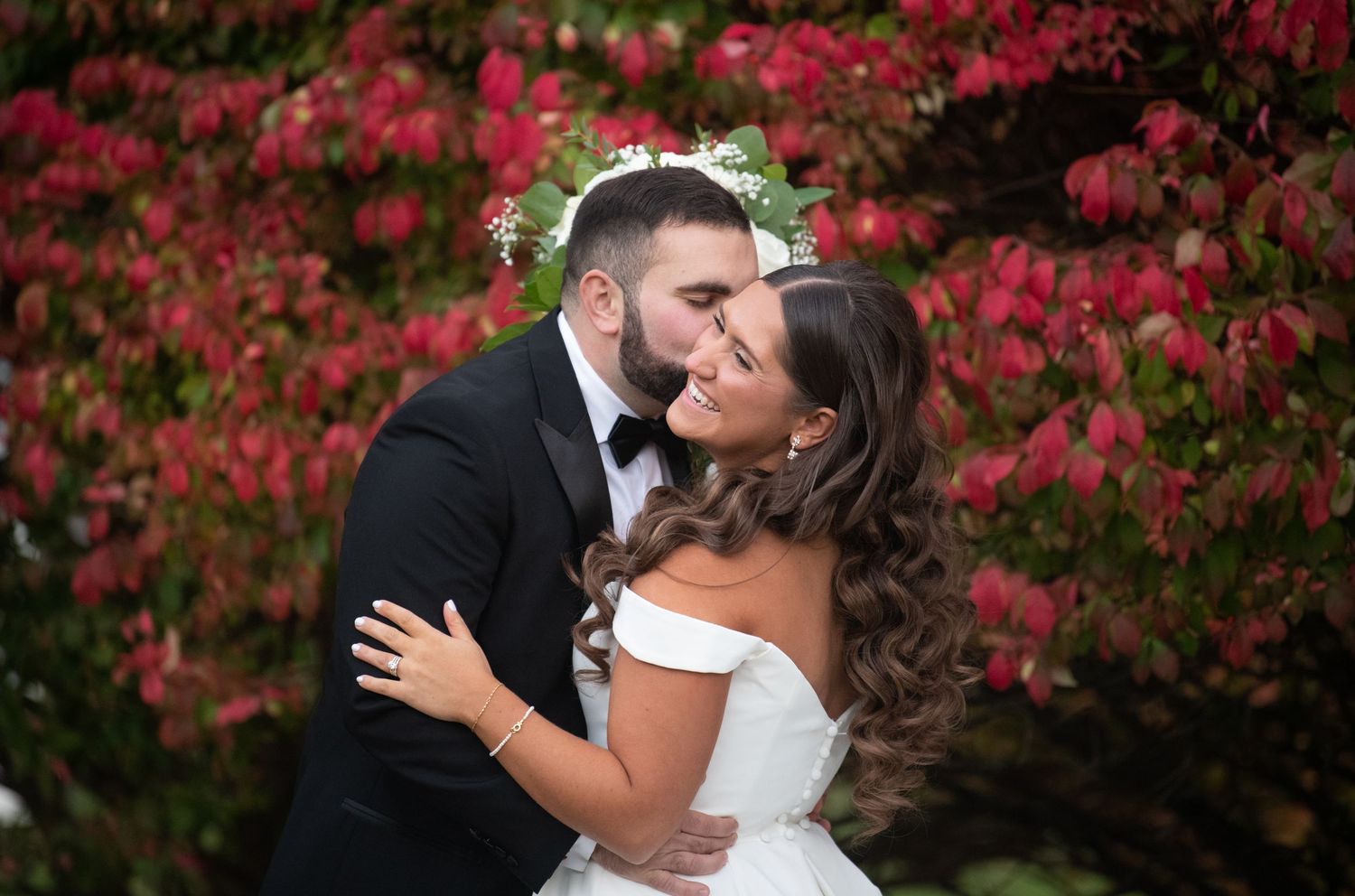 A romantic wedding portrait of a couple embracing against vibrant red autumn foliage in a garden setting.
