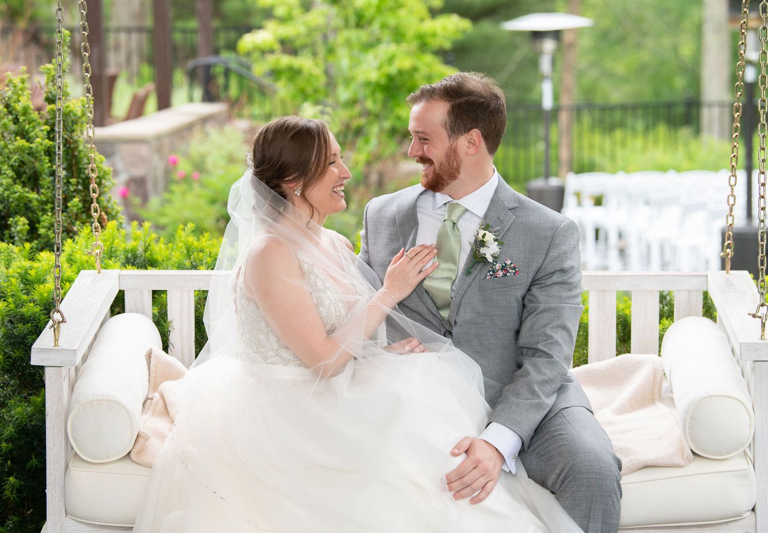 A couple shares an intimate moment on a white porch swing surrounded by lush garden greenery and wedding decor.