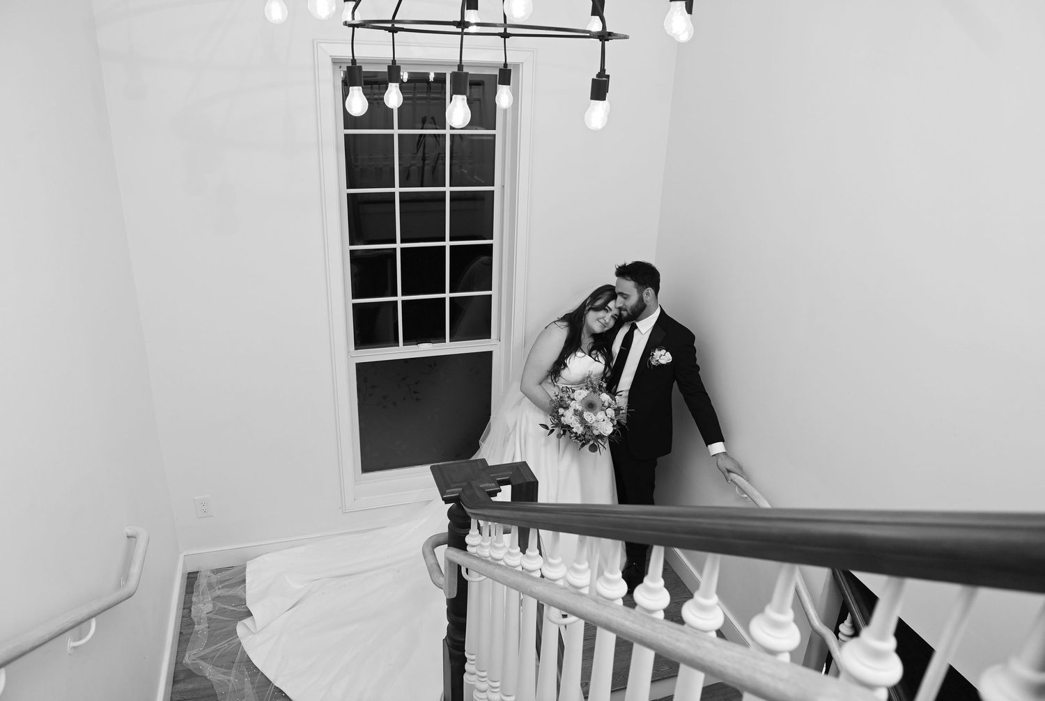 A black and white photograph captures a romantic moment on a staircase with elegant lighting and window details.