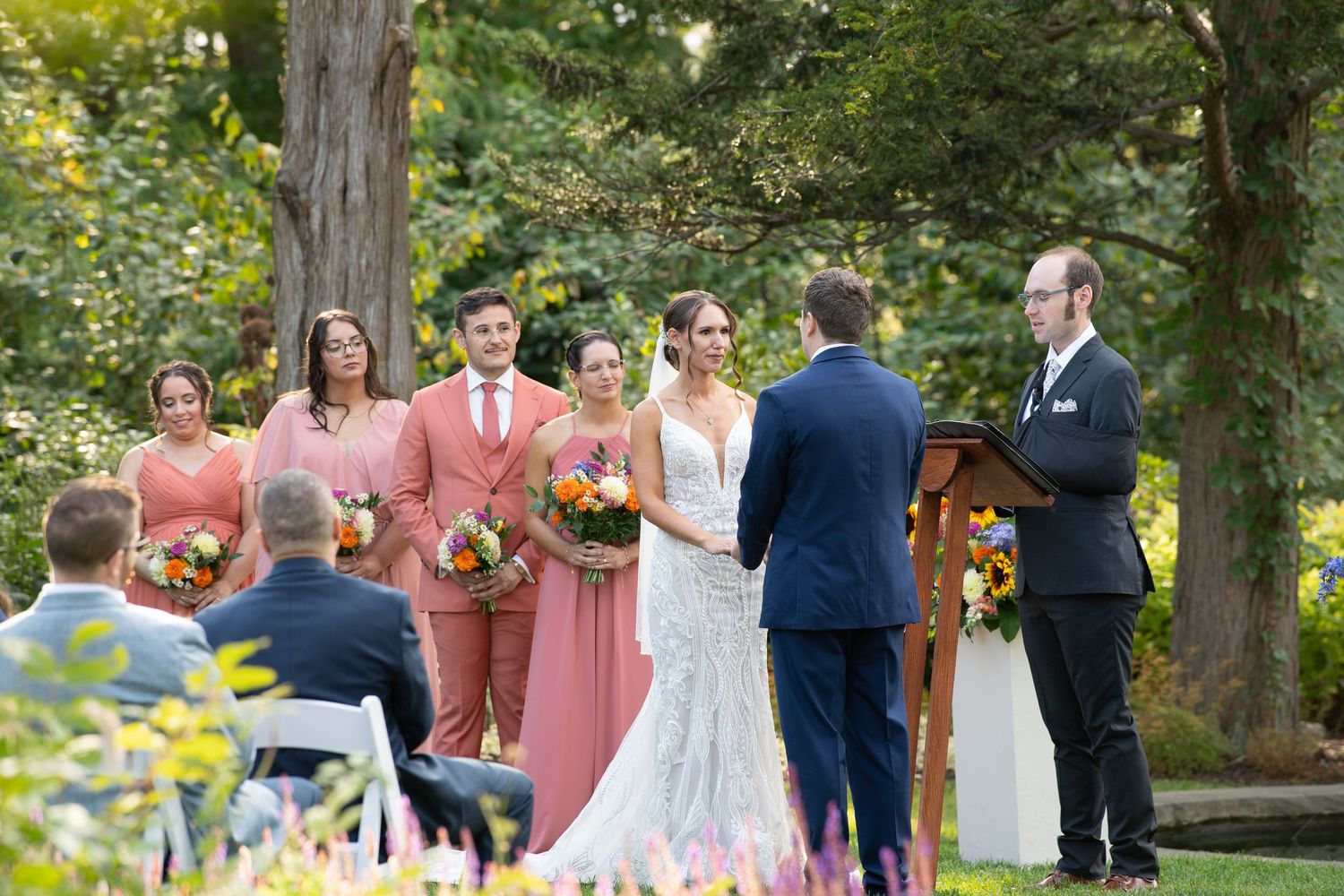 Wedding ceremony takes place under tall trees in a lush garden setting with guests in coral and navy attire.