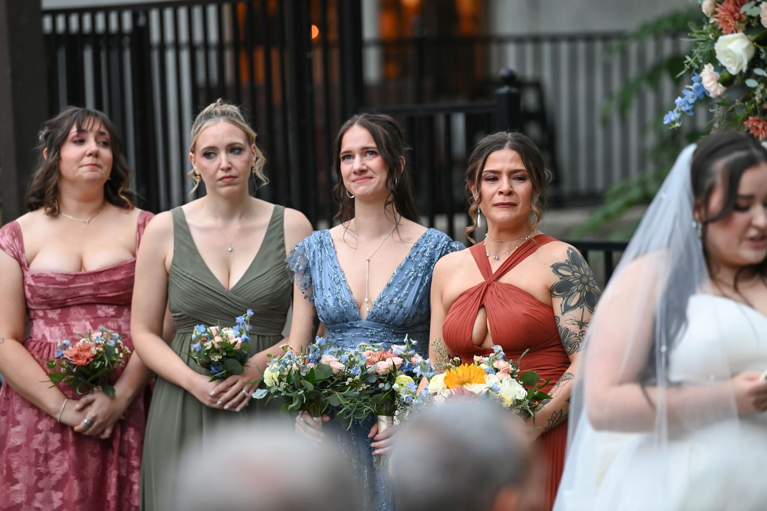A group of bridesmaids in varying shades of pink and blue dresses pose together at an outdoor wedding.