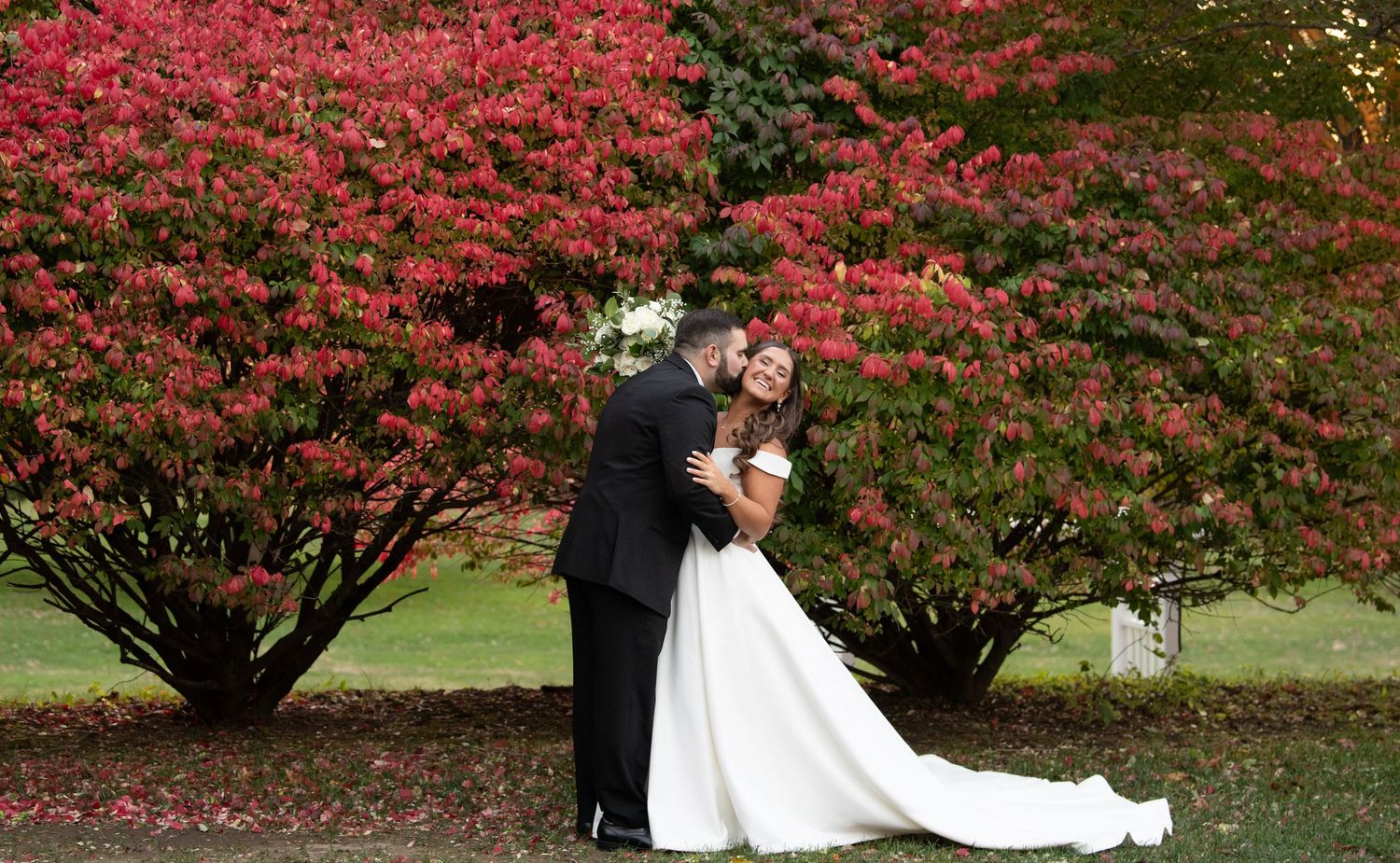 A couple embraces in formal wedding attire against vibrant red autumn foliage in a garden setting.
