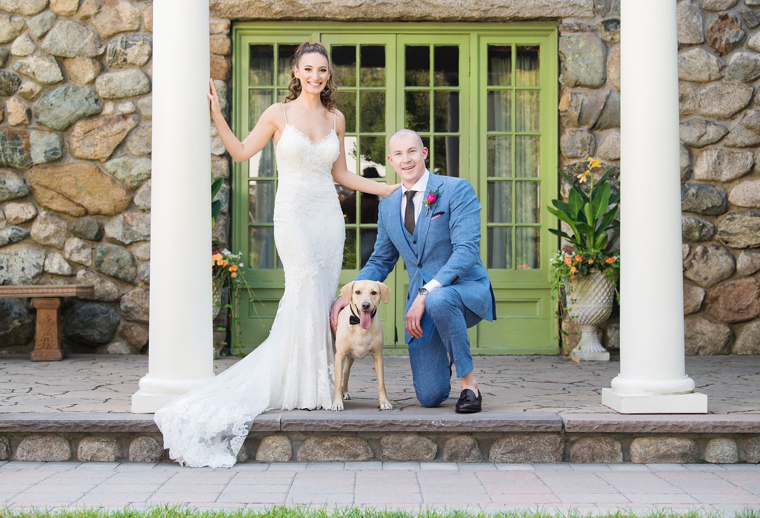 A wedding couple poses with their golden retriever on stone steps by a green door entrance with rustic stone walls.