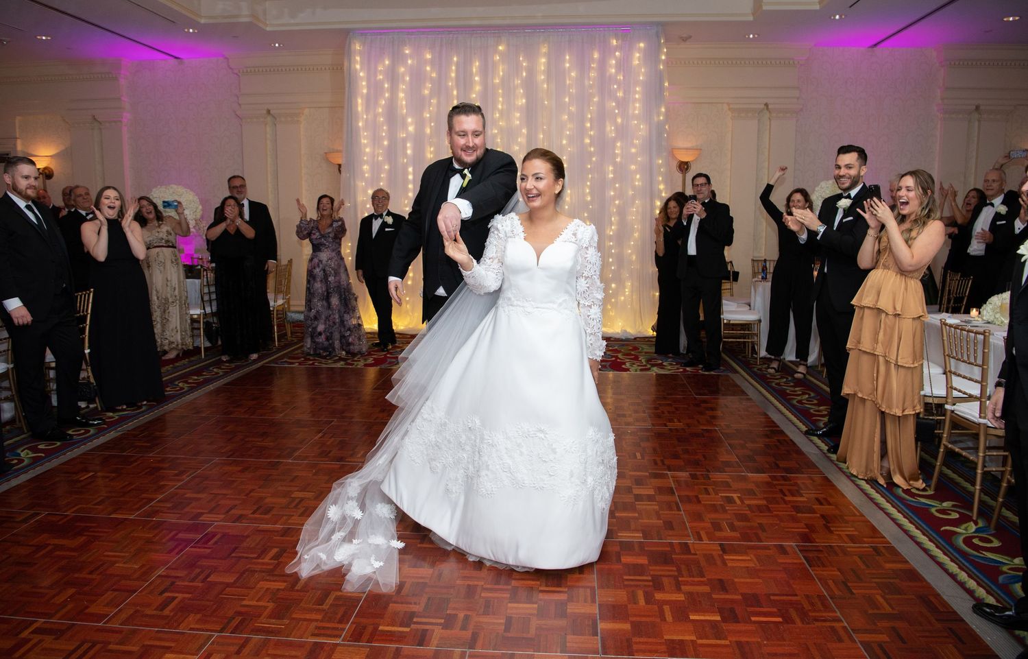 Wedding couple dances their first dance on a wooden floor under purple lighting while guests watch from the sides.