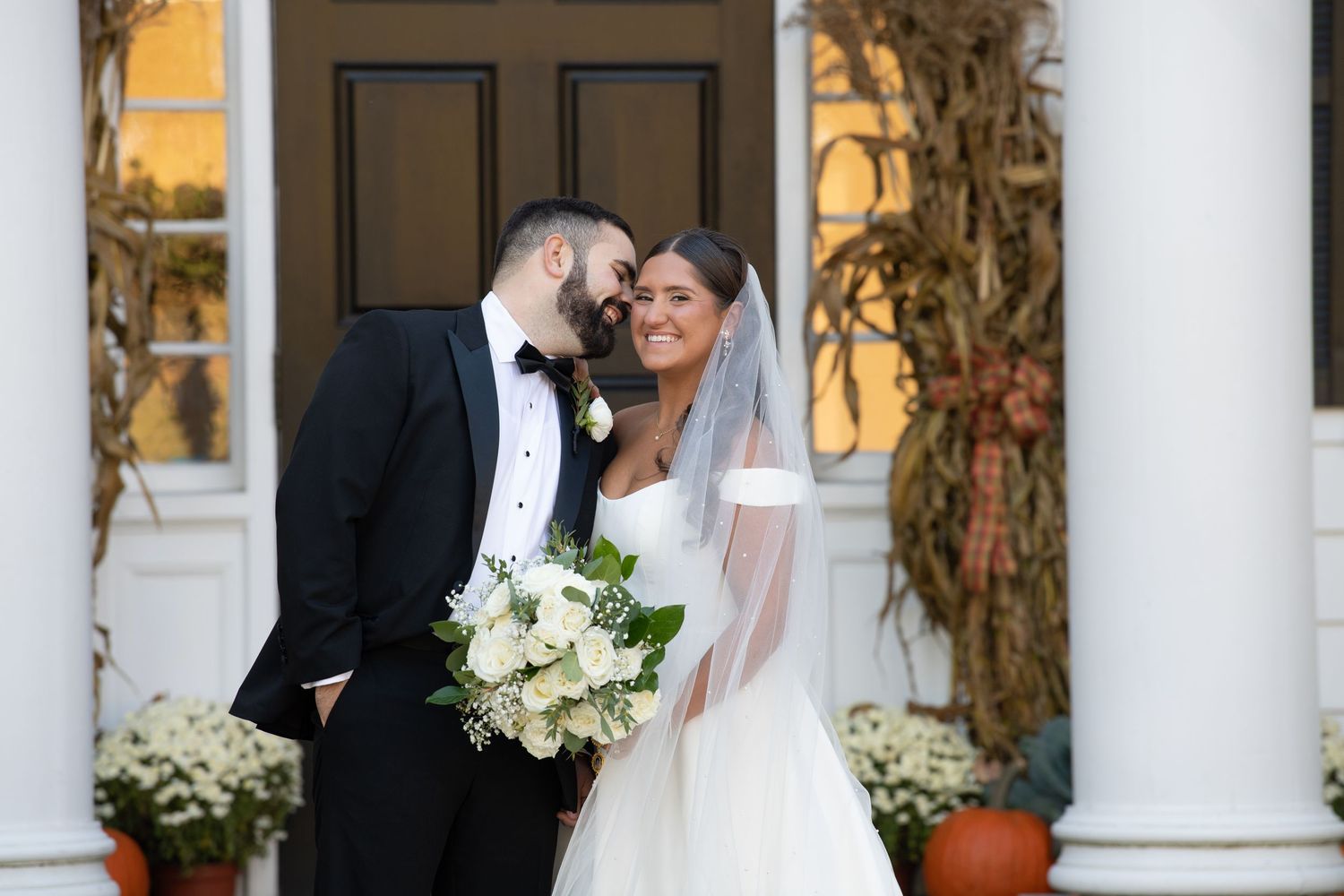 A wedding couple shares an intimate moment on a white-columned porch decorated with floral arrangements at sunset.