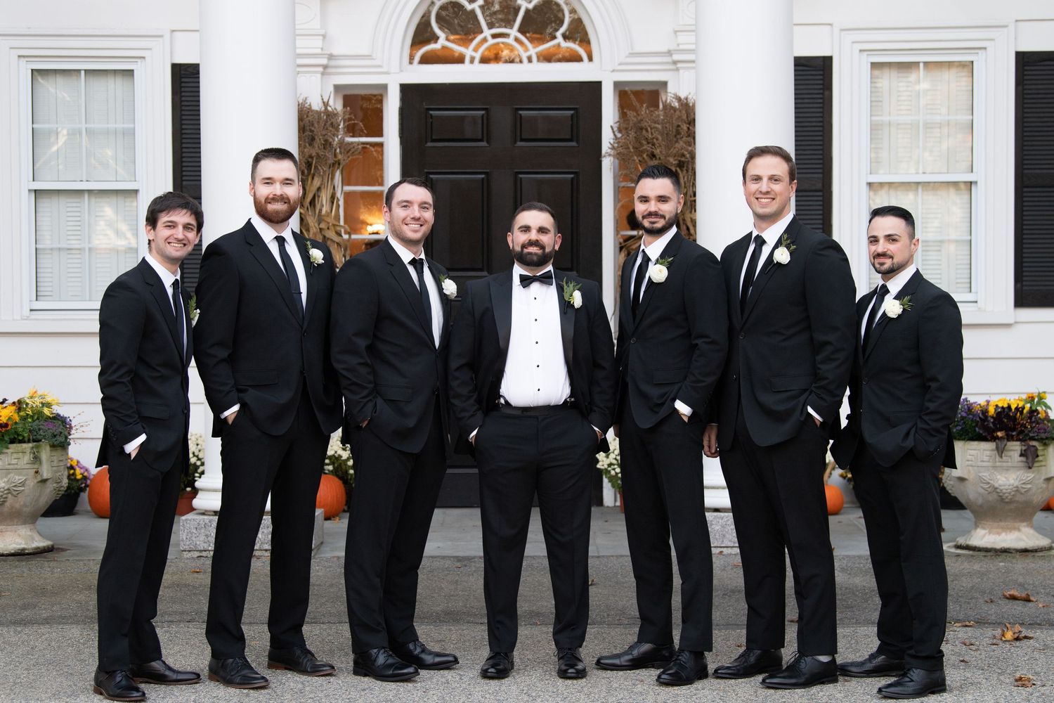 A group of seven men in black tuxedos and bow ties stand together smiling in front of a white building entrance.