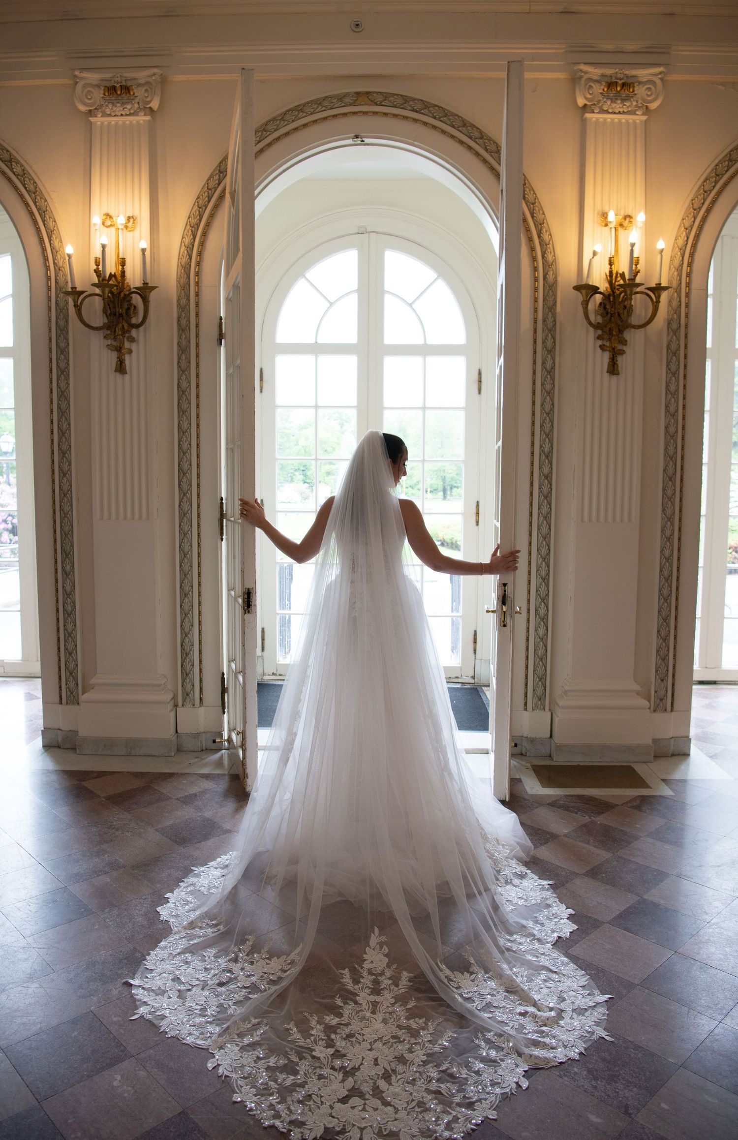 Bride in a lace-trimmed cathedral veil stands at ornate French doors in an elegant ballroom.