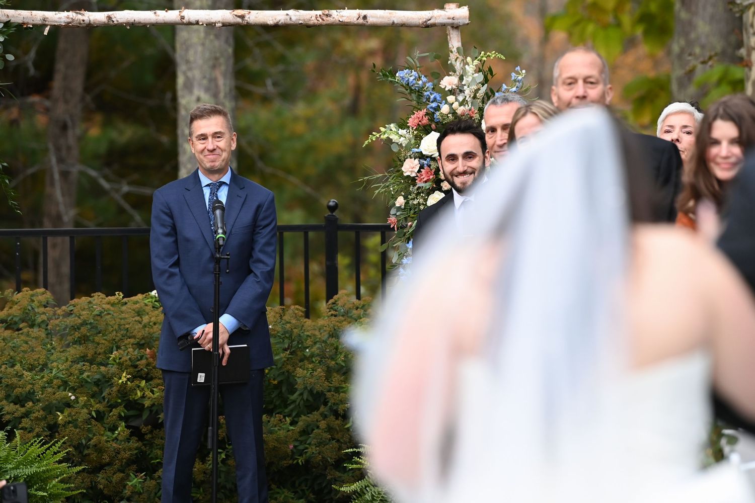 A couple poses together outdoors during a fall wedding ceremony with colorful autumn foliage in the background.
