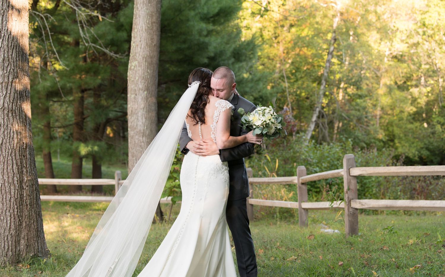 A bride and groom share a romantic kiss in a wooded outdoor setting with sunlight filtering through the trees.