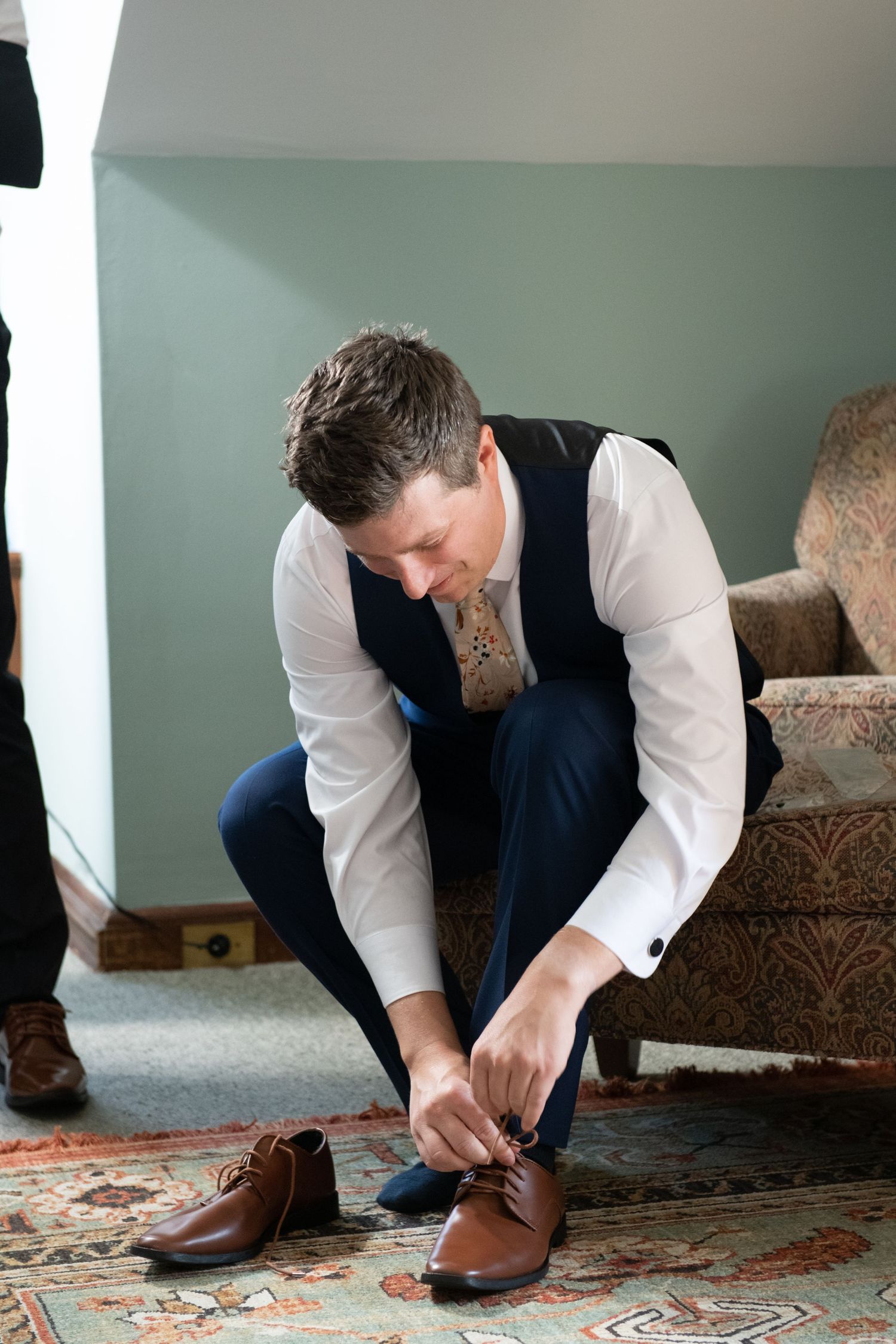 Person in formal attire tying brown leather dress shoes while sitting on patterned chair.