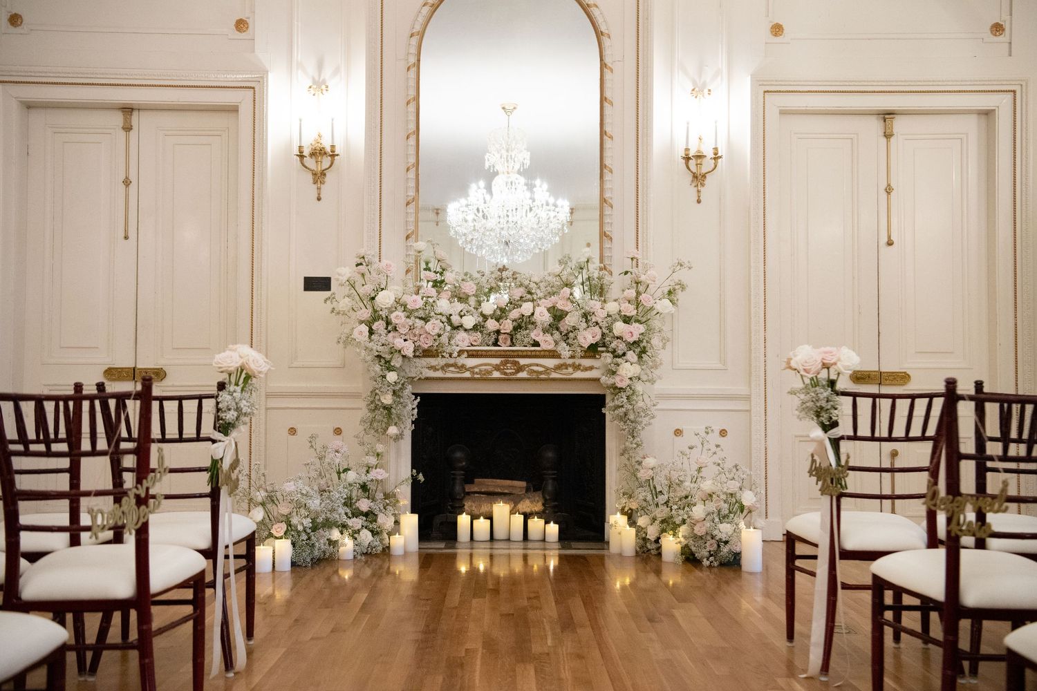 Elegant wedding ceremony setup with floral arch over fireplace, chandelier, candles, and chiavari chairs in a classic ballroom.