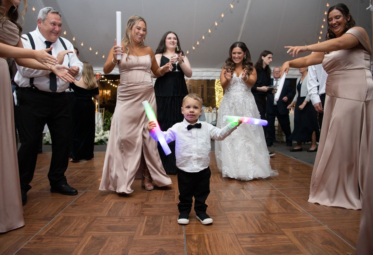Young ring bearer in a bow tie dances enthusiastically on wooden dance floor at wedding reception while guests smile and cheer.